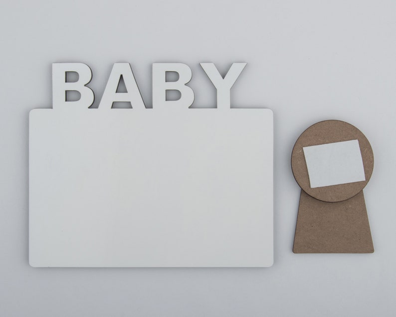 May include: A white wooden photo frame with the word "BABY" in capital letters above the frame. A separate, round, brown wooden piece with a white square is next to the frame.