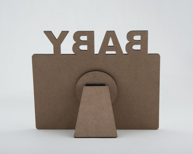 May include: A brown wooden picture frame with the word "BABY" cut out at the top. The frame has a rectangular shape with a circular support and a triangular stand on the back.