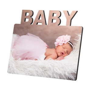 May include: A wooden photo frame with the word "BABY" at the top. The frame displays a photo of a sleeping infant wearing a pink tutu and a flower headband, resting on a fluffy white surface.