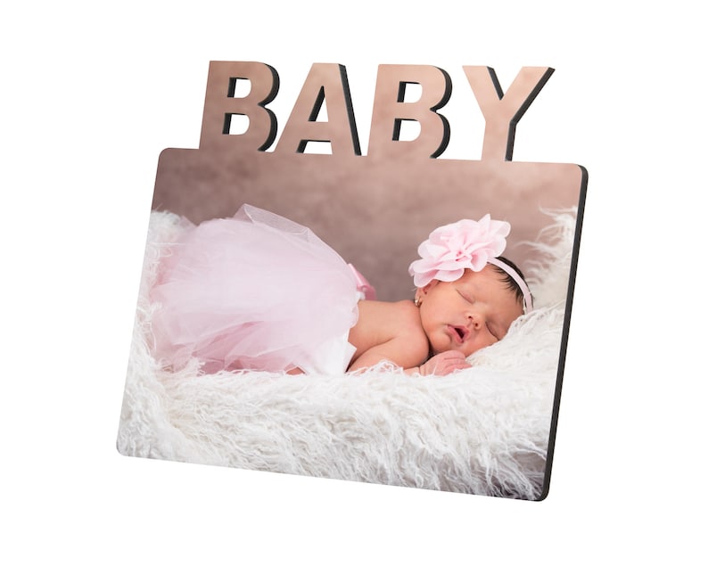 May include: A wooden photo frame with the word "BABY" at the top. The frame displays a photo of a sleeping infant wearing a pink tutu and a flower headband, resting on a fluffy white surface.