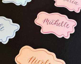 May include: A collection of die-cut name tags with wavy, cloud-like edges in pastel shades of light blue, pink, and peach. Each tag features a handwritten-style script font displaying names including Kayla, Maria, Michelle, and Nicole.