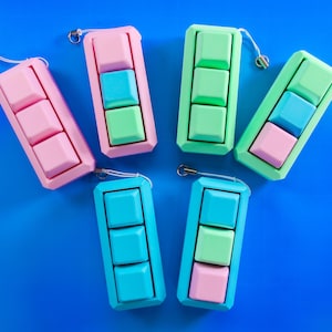 Pode incluir: Seis brinquedos fidget coloridos em rosa, verde e azul estão dispostos sobre uma superfície azul. Cada brinquedo tem três botões quadrados. Os brinquedos têm um pequeno laço de metal preso a um cordão branco.