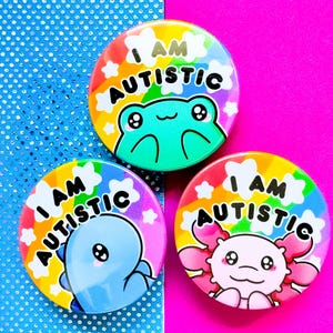 Può includere: Tre bottoni rotondi colorati con la scritta "I AM AUTISTIC". Ogni bottone presenta un diverso animale dei cartoni animati: una rana, un dinosauro e un axolotl. I bottoni hanno uno sfondo arcobaleno.