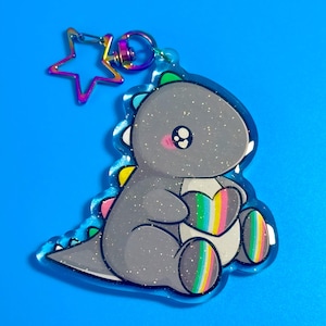Pode incluir: Um porta-chaves de acrílico transparente com um dinossauro cinzento brilhante que segura um coração com riscas arco-íris. O dinossauro tem uma barriga branca, bochechas cor-de-rosa e um fecho em forma de estrela com acabamento arco-íris iridescente.