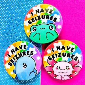 Könnte beinhalten: Drei bunte runde Buttons mit dem Text "I HAVE SEIZURES". Jeder Button zeigt ein anderes Zeichentier: einen Frosch, einen Dinosaurier und einen Axolotl. Die Buttons haben einen Regenbogenhintergrund und weiße Sternakzente.