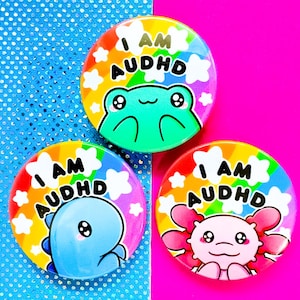 Può includere: Tre bottoni rotondi colorati con la scritta "I AM AUDHD". Ogni bottone presenta un diverso animale dei cartoni animati: una rana, un dinosauro e un axolotl. I bottoni hanno uno sfondo arcobaleno e accenti di stelle bianche.