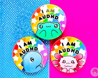 Könnte beinhalten: Drei bunte runde Buttons mit dem Text "I AM AUDHD". Jeder Button zeigt ein anderes Zeichentier: einen Frosch, einen Dinosaurier und einen Axolotl. Die Buttons haben einen Regenbogenhintergrund und weiße Sternakzente.