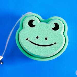 Op de afbeelding: Een groene, intrekbare sleutelhanger in de vorm van een kikker met zwarte ogen en een glimlachende mond.