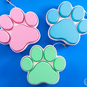 Może przedstawiać: Trzy kolorowe breloczki w kształcie łapy z białym obramowaniem. Breloczki są różowe, niebieskie i zielone.