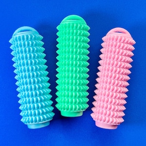 Op de afbeelding: Drie kleurrijke hondenspeeltjes in blauw, groen en roze, geplaatst op een blauwe achtergrond. Elk speeltje is cilindrisch met een getextureerd, geribbeld oppervlak en een lus aan de bovenkant. Deze kauwspeeltjes zijn ontworpen voor huisdieren.