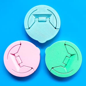 May include: Three round, colorful phone grips in pastel shades of blue, pink, and green are displayed against a bright blue background. Each grip has a unique design with a hinged, folding mechanism for secure phone handling.