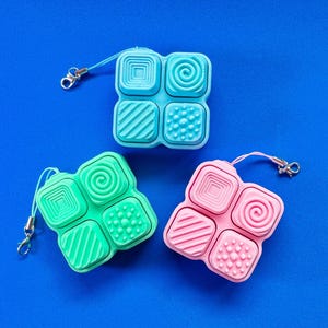 May include: Three colorful, square fidget toys with textured surfaces and attached lanyards. The toys are in shades of blue, green, and pink, each with a different pattern on the four squares. The background is a solid blue.