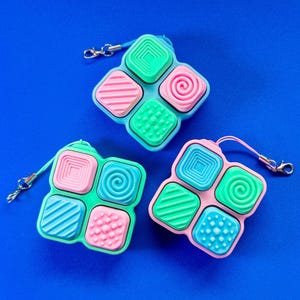 Puede incluir: Tres juguetes fidget coloridos con cuadrados texturizados en rosa, verde y azul. Cada juguete tiene un patrón diferente en los cuadrados, incluyendo líneas, puntos y espirales. Tienen un cordón pequeño y un cierre de metal.