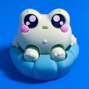 May include: A pale green frog figurine with large black eyes, white highlights, and pink circular cheeks. The frog is seated inside a textured blue circular base. The figure has a smooth, matte finish and a rounded, symmetrical appearance.
