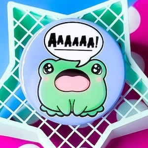 Peut inclure: Un bouton bleu et vert avec une grenouille de dessin animé aux grands yeux et une bouche ouverte disant "AAAAAAA!" dans une bulle de dialogue.