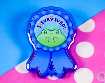 May include: A blue ribbon-shaped badge with a cartoon frog illustration. The badge has the text "I SURVIVED!" in a circle around the frog. The frog is green with a smiling face. The background has colorful sprinkles.