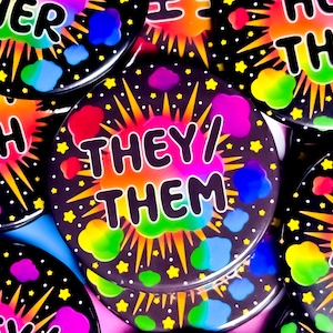 Op de afbeelding: Een verzameling zwarte ronde buttons met de tekst "THEY/ THEM" in het wit. Elke button heeft een kleurrijk, explosie-achtig ontwerp met regenboogkleuren en gele sterren.