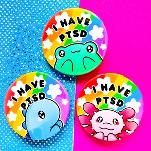 Può includere: Tre spille arcobaleno con il testo "I Have PTSD" e un animale dei cartoni animati su ciascuna. Gli animali sono una rana, un axolotl blu e un axolotl rosa.