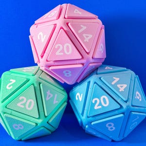 May include: Three colorful, geometric D20 dice in pink, green, and blue, each with white numbers. The dice are arranged against a solid blue background, showcasing their triangular facets and numerical markings.