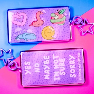 May include: Two purple acrylic keychains with a star-shaped charm. The top keychain features a pink heart, a red worm, a yellow worm, and a green apple with a worm. The bottom keychain has the words "YES NO MAYBE I'M NOT SURE SORRY" printed on it.