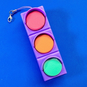 May include: A purple, handheld traffic light toy with three circular lights in red, orange, and green. The toy has a small silver clasp and a purple lanyard attached to the top. The background is a solid blue.