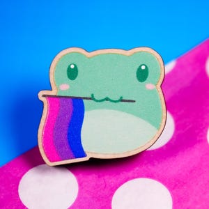 May include: A wooden frog-shaped pin with a light green body, pink cheeks, and large green eyes. The frog holds a flag with the colors of the bisexual pride flag: pink, purple, and blue. The pin is set against a blue and pink polka dot background.