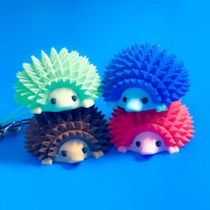May include: Four small hedgehog figurines stacked in two pairs. The top left is light green, the bottom left is dark brown, the top right is blue, and the bottom right is red with a pink face. A silver metal keychain attachment is on the left.