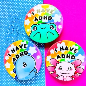 Może przedstawiać: Trzy okrągłe guziki z napisem "I HAVE ADHD". Każdy guzik przedstawia inne zwierzę z kreskówki: żabę, niebieskie stworzenie podobne do dinozaura i różowego aksolotla. Tła są w kolorach tęczy.