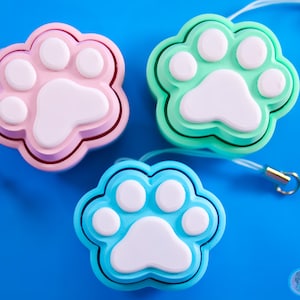 Może przedstawiać: Trzy breloki w kształcie łapy w kolorze różowym, jasnozielonym i jasnoniebieskim. Każdy brelok ma biały odcisk łapy w środku.