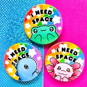 May include: Three colorful round buttons with the text "I NEED SPACE". Each button features a different cartoon animal: a frog, a blue dinosaur, and a pink axolotl. The buttons have a rainbow background with white clouds.
