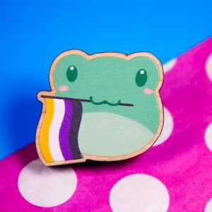 May include: A green frog with a big smile holding a yellow, white and purple pride flag. The frog is made of wood and has a cartoonish style.