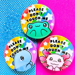 Puede incluir: Tres botones redondos de colores arcoíris con el texto "Please Don't Touch Me" y un animal de dibujos animados en cada uno. Los animales son una rana, una axolotl azul y una axolotl rosa.