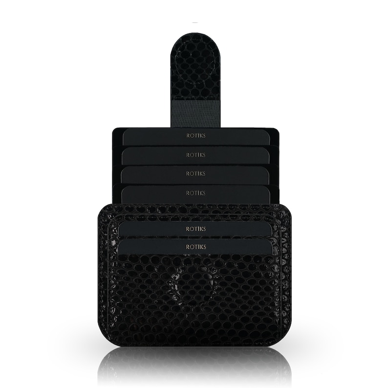 May include: Black leather card holder with a textured, crocodile-like pattern. The holder has a flap with a snap closure and multiple slots for cards. The text "ROTIKS" is embossed on the cards.