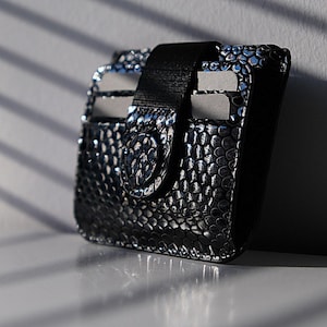 May include: A black leather card holder with a snakeskin pattern and a black elastic band closure.