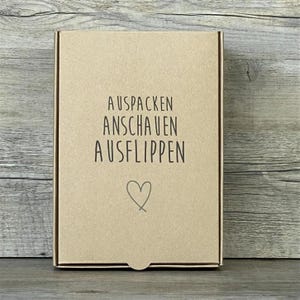Könnte beinhalten: Ein brauner Karton mit dem Text "AUSPACKEN ANSCHAUEN AUSFLIPPEN" und einem Herzsymbol.