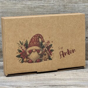 Könnte beinhalten: Eine braune Pappschachtel mit einer festlichen Zwergenillustration. Der Zwerg trägt einen rot-weißen Hut und hat einen langen weißen Bart. Die Schachtel zeigt auch ein kleines Haus und den Text "Für Anton" in Burgunderrot, ideal zum Verschenken.