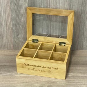Könnte beinhalten: Eine Teebox aus Bambus mit einem Deckel aus klarem Glas und sechs Fächern. Der Text "Trink einen Tee. lies ein Buch, mach's dir gemütlich" ist auf der Vorderseite eingraviert. Der Deckel ist mit Scharnieren versehen.