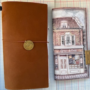Könnte beinhalten: Ein braunes Leder-Notizbuch mit einem Messing-Charm und elastischem Verschluss. Daneben befindet sich eine Notizbucheinlage mit einer Illustration des Gran Cafe-Geschäfts. Ein kleines Messingschild mit Text ist ebenfalls sichtbar.