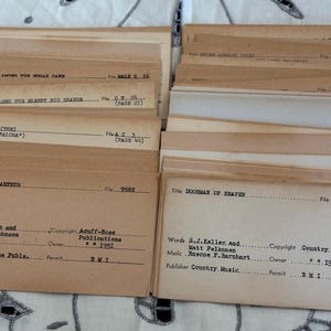 Może przedstawiać: Kolekcja zabytkowych kart indeksowych, prawdopodobnie do muzyki lub tekstów piosenek, ze szczegółami pisanymi ręcznie i na maszynie. Karty są beżowe i brązowe, z tytułami i informacjami takimi jak "DOORMAN OF HEAVEN" i "DOUG MacARTHUR".