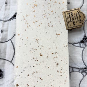 Puede incluir: Un cuaderno rectangular con una cubierta texturizada de color blanco roto con copos de pan de oro esparcidos. Un clip de metal color latón está sujeto en la esquina superior derecha, grabado con el texto TRAVELER'S COMPANY JAPAN TRAVELER'S notebook MADE IN JAPAN.