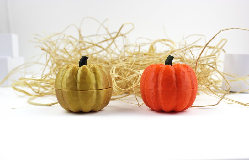 May include: Two small pumpkin-shaped containers, one gold and one orange, are sitting on a white surface. The containers are surrounded by straw.