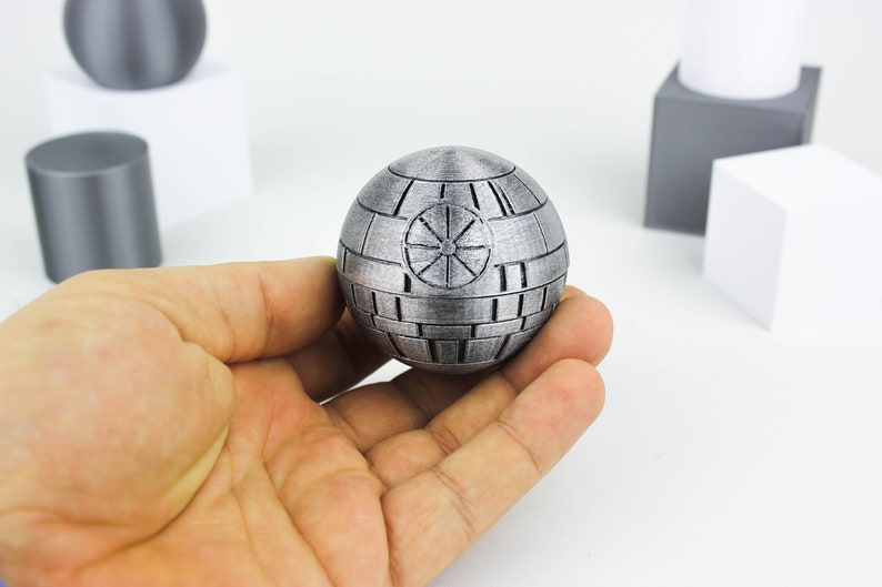 Puede incluir: Una r&eacute;plica de la Estrella de la Muerte de color plateado, un objeto de colecci&oacute;n popular de Star Wars, se sostiene en la mano de una persona. La r&eacute;plica est&aacute; detallada con las trincheras ic&oacute;nicas y un panel central.