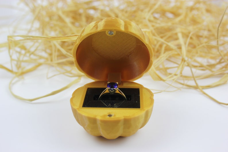 May include: A gold ring with a purple gemstone sits inside a yellow pumpkin-shaped ring box.