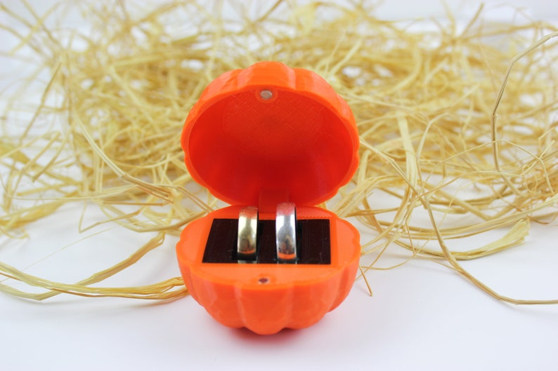 May include: An orange pumpkin-shaped ring box with the lid open, revealing two silver rings inside.