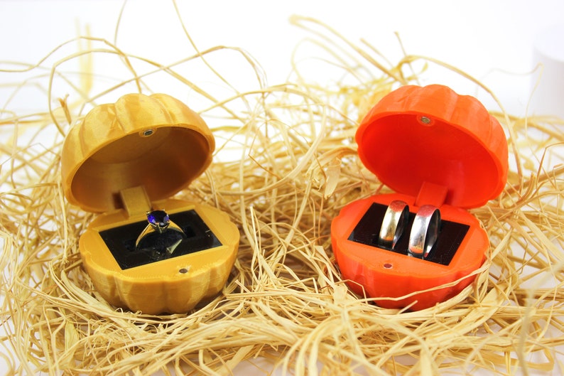May include: Two pumpkin-shaped ring boxes, one orange and one yellow, are open and sitting on a bed of straw. The orange box contains two silver wedding bands, and the yellow box contains a single ring with a blue gemstone.