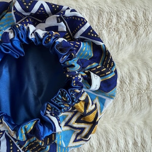 Peut inclure: Bonnet doublé en satin bleu et or avec un motif géométrique. Le bonnet a une fermeture à cordon et une étiquette qui indique "Cozy Co."
