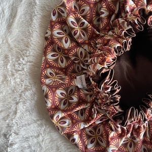 Peut inclure: Un bonnet de nuit en satin à motif marron, blanc et orange avec une doublure en satin marron. Le bonnet a une fermeture à cordon et le texte "Housewife" est imprimé sur le cordon.