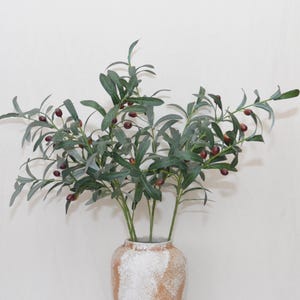 May include: An arrangement of olive branches with green leaves and small, dark red olives, displayed in a textured, light brown and white vase. The branches create a natural and rustic aesthetic, suitable for home decor.