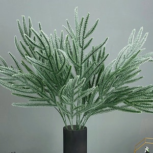 May include: A bunch of artificial green pine branches with a white frosted finish. The branches are arranged in a vase.
