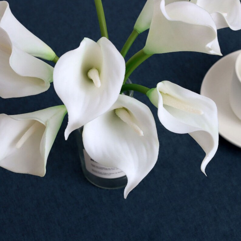 May include: A bouquet of white calla lilies arranged in a clear glass vase. The lilies are in full bloom and have a delicate, elegant appearance.